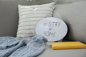 Book, sweater and speech bubble with hashtag STAY AT HOME on sofa, closeup. Message to promote self-isolation during COVIDÃ¢â¬â19