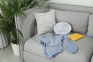 Book, sweater and speech bubble with hashtag STAY AT HOME on sofa indoors. Message to promote self-isolation during COVIDÃ¢â¬â19