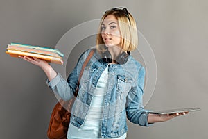 Book vs. e-book. Girl student holding a traditional textbook and reading e-books isolated on gray background. Choose between paper
