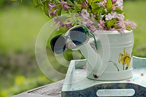 Book on the white vintage table with vase and sunglasses on green grass on sunny summer day, soft focus. Lawn with spring flowers
