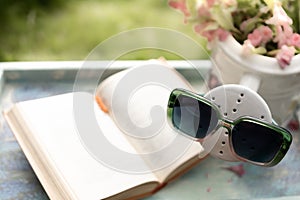 Book on the white vintage table with vase and sunglasses on green grass on sunny summer day, soft focus. Lawn with spring flowers