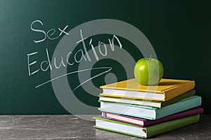 Books and apple on grey table near chalkboard with phrase `Sex education`