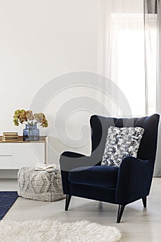 Books and flowers on console table in white living room interior with velvet wing back chair with patterned pillow