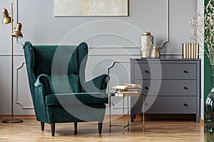 Books on stylish golden small table next to emerald green velvet wing back chair in grey living room interior