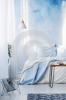 Books on table and white and blue bedding on bed in pastel bedroom interior with lamp