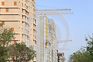 Booms of construction cranes among residential buildings under construction among thickets of trees