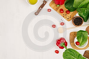 Border of fresh green greens, red paprika, cherry tomato, pepper, oil and utensils on soft white wooden background.