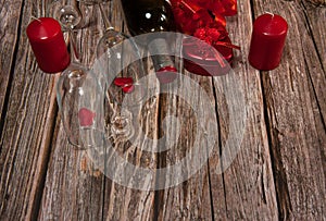 A border of two narrow empty glasses, a gift box, candles, red hearts and a bottle of wine on a wooden background.