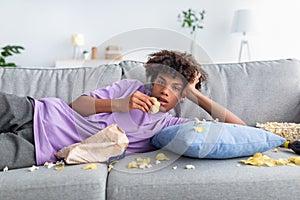 Bored Afro teenager lying on sofa with scattered snacks, eating chips, watching dull show or movie on TV at home