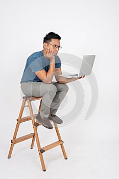 A bored Asian man is waiting for an email on his laptop while sitting on a wooden ladder. isolated
