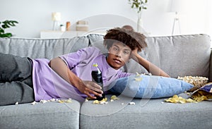 Bored black teenager lying on couch with scattered food, watching dull show or movie on TV, killing time at home