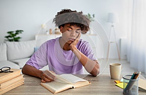 Bored black teenager sitting at desk, reading book, studying at home