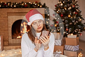 Bored Caucasian woman wearing white sweater and santa claus hat using smart phone, scrolling online with calm expression, sitting