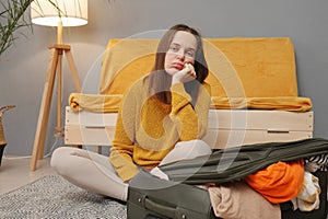 Bored Caucasian young adult woman packing suitcase at home taking too much attire waiting for her departure with sad boring facial