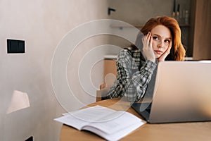 Bored female student studying using laptop sitting at desk with book in home. Exhausted woman cannot concentrate on