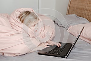 Bored girl lying in bed under blanket and learning in virtual online school or class. Kid working on laptop Internet at home.