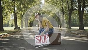 Bored hippie retro woman sitting on suitcase with Stop banner. Portrait of young Caucasian woman hitchhiking on sunny