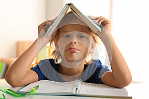 Bored little boy with book on his head doing homework at table indoors