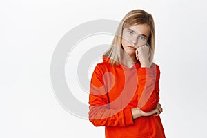 Bored little girl looking with sad, gloomy and unamused face at camera, standing upset and moody against white