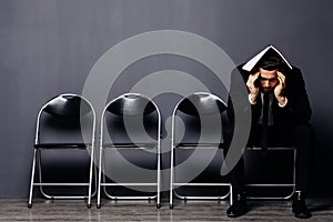 Bored young man in formal suit sitting on office chair in waiting room with document or resume in hand. Young job candidate or bus