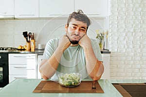 A bored man sits in the kitchen with his head in his hands in front of a plate of salad and a fork.