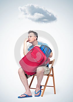 Bored man in summer clothes with a red suitcase sitting on a chair waiting. Over the head of a cloud