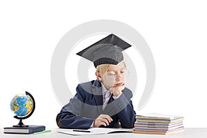 Bored schoolboy does his homework. Boy in school uniform and student hat sits at table next to notebooks and globe