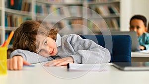 Bored schoolboy lying on table in library or classroom during uninterested lesson, looking away and thinking