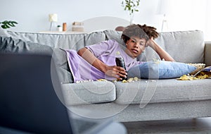 Bored teen guy lying on sofa with scattered snacks, watching movie on laptop at home