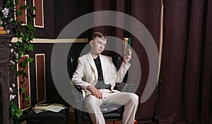 Bored teen student guy in white business suit sitting in a chair with a book by the fireplace