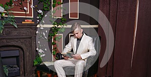 Bored teen student guy in white business suit sitting in a chair with a book by the fireplace