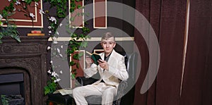 Bored teen student guy in white business suit sitting in a chair with a book by the fireplace