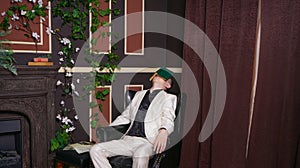 Bored teen student guy in white business suit sitting in a chair with a book by the fireplace