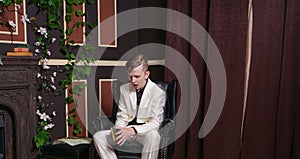 Bored teen student guy in white business suit sitting in a chair with a book by the fireplace