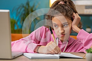 Bored tired school girl doing homework exercise online, typing on laptop makes notes in notebook