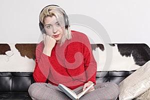 Bored woman with headphones sitting on leather couch holding book