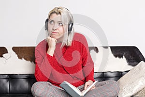 Bored woman with headphones sitting on leather couch holding book