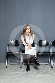 Bored young businesswoman holding paper and looking away while sitting on chair against grey background