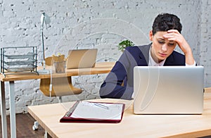 Bored young businesswoman sitting with laptop at desk against brick wall in office