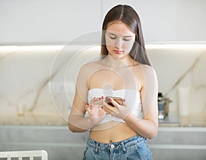 Bored young woman stands in kitchen leans on countertop and surfs internet. Home pastime