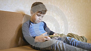 Boring boy uses cellphone while sitting alone at home on the couch. Caucasian preschooler boy looking at phone screen