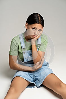 Boring brunette tan teen girl sitting on gray background looking at camera resting cheek on hand.