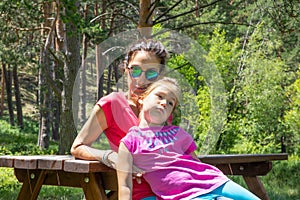 Boring little girl resting sitting on her mother on a picnic bench in a forest of the country