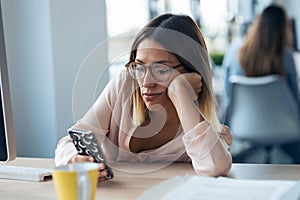 Boring young business woman sending messages with mobile phone while sitting in modern startup office