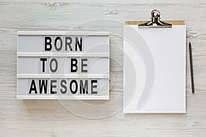 `Born to be awesome` words on a lightbox, clipboard with blank sheet of paper on a white wooden surface, top view. Overhead, fro