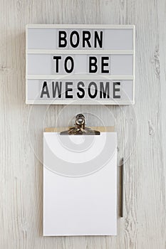 `Born to be awesome` words on a modern board, clipboard with blank sheet of paper on a white wooden background, top view. Overhe