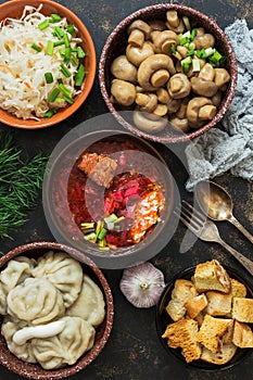 Borsch, dumplings, sauerkraut, pickled mushrooms. Russian dishes on a dark rustic background, top view. Traditional Russian cuisin