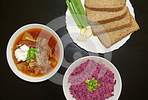 Borscht soup and beet salad in a white bowls with black bread, green onion and garlic on dark wooden table.