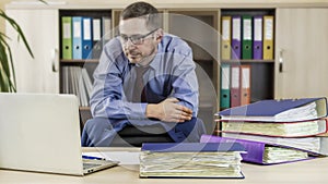 Boss businessman at his desk in the office with a bunch of folders. Leans against the back of the chair and looks at the laptop.