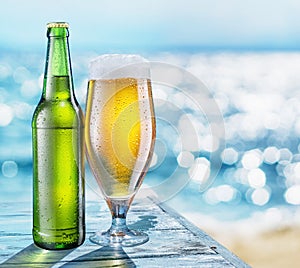 Bottle of beer and beer glass with condensation on the wooden table. Blurred sparkling sea at the background. Summer vibes